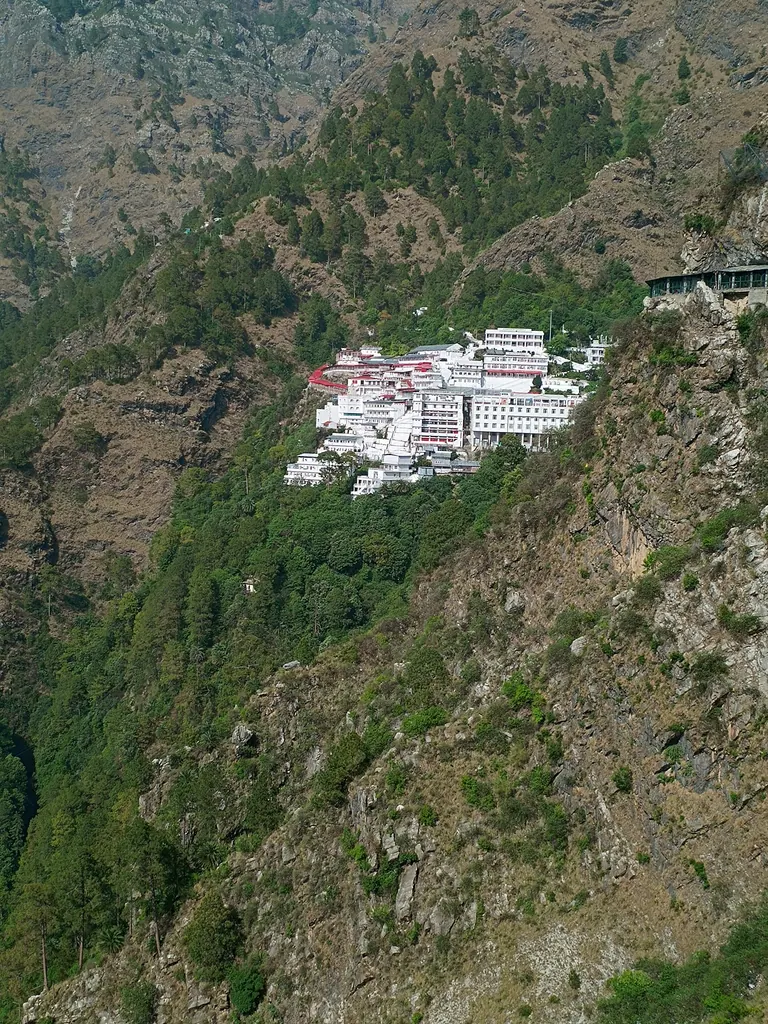 The Vaishno Devi Bhawan complex seen across a green forested slope from the trekking track