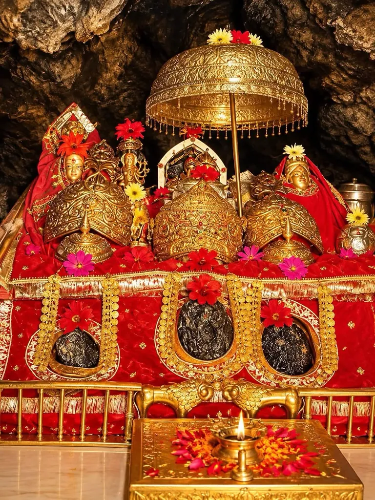 The three Holy Pindis of Mata Vaishno Devi in the cave sanctum, adorned with red cloth, flowers and golden crowns beneath a golden canopy, with a lit lamp in front