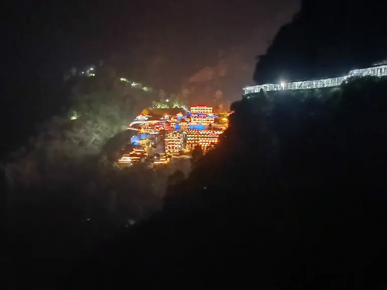 The Vaishno Devi Bhawan complex illuminated at night against dark mountains, with the lit track running along the slope