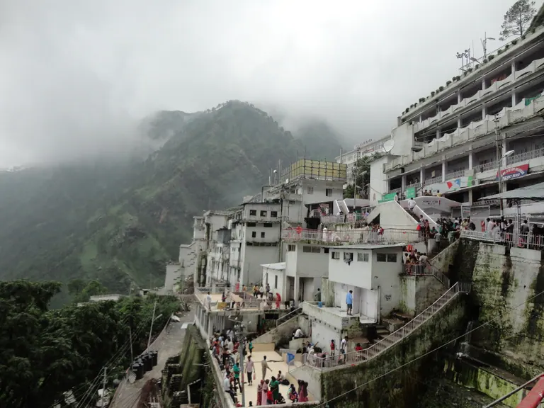 Multi-storey buildings of the Vaishno Devi Bhawan complex built into the mountainside with clouds drifting over the hills