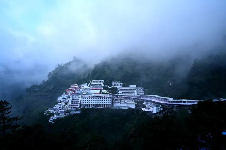 The white buildings of the Mata Vaishno Devi Bhawan on the Trikuta hillside with fog rising from the forested valley